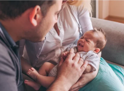 Une maman est assise sur son canapé. Elle tient son nourrisson de quelques jours dans les bras, sur ses genoux. le papa à côté d'elle se penche sur le bébé. le bébé semble en train de pleurer.