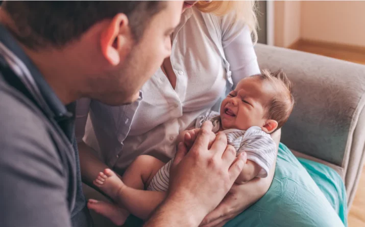 Une maman est assise sur son canapé. Elle tient son nourrisson de quelques jours dans les bras, sur ses genoux. le papa à côté d'elle se penche sur le bébé. le bébé semble en train de pleurer.